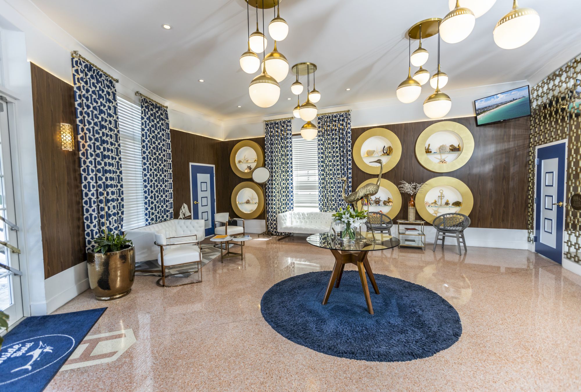 A modern lobby with white seating, blue accents, round shelves, geometric curtains, and elegant lighting. A sculpture adorns a glass table.