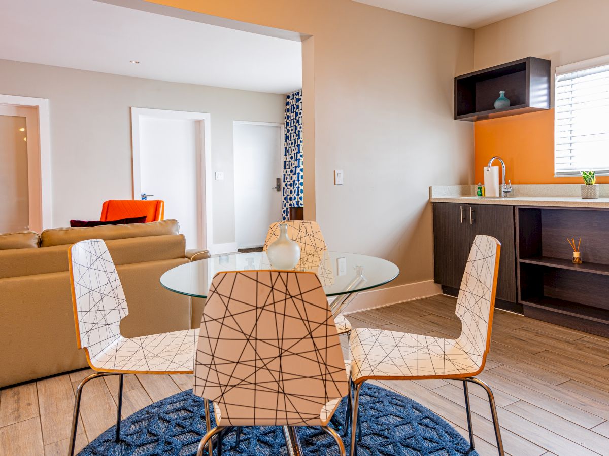 Modern living area with a round glass table, patterned chairs, couch, counter with a sink, and shelves. A blue rug accentuates the space.