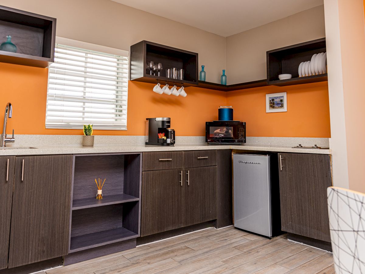 A kitchenette with orange walls, cabinets, microwave, mini-fridge, coffee maker, sink, cups, and decor items.