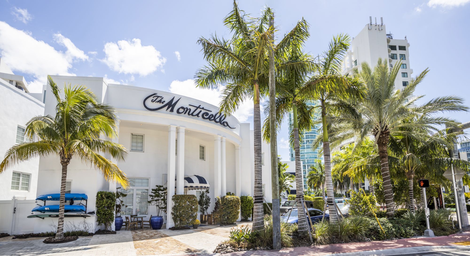A white building named "The Monticello" surrounded by palm trees and greenery with a few canoes to the side.