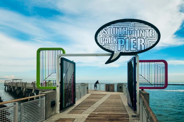 A pier entrance with a sign reading "South Pointe Park Pier" against a bright blue sky, arched over a wooden path extending into the water.