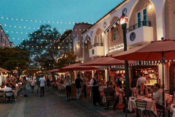 A bustling outdoor street scene features diners at tables under umbrellas, surrounded by string lights and a lively atmosphere.