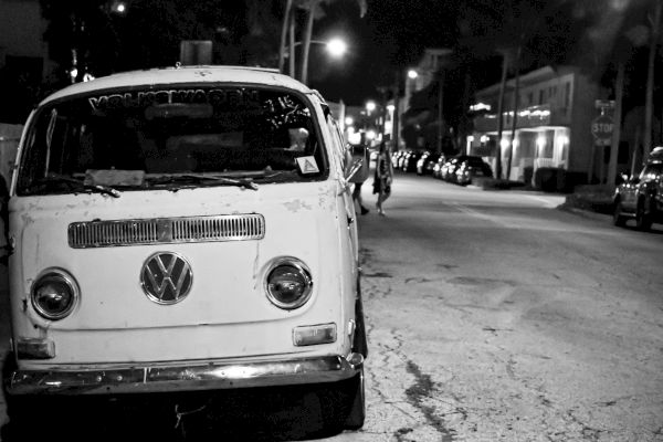 A black and white image of a vintage Volkswagen van parked on a dimly lit street at night with palm trees and buildings in the background.