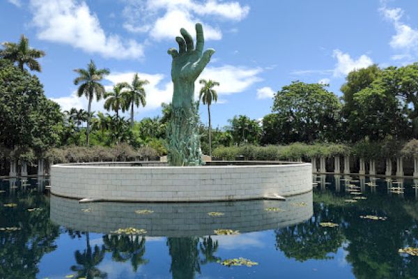 A sculpture of a large hand reaching upward is surrounded by water, with trees and a clear blue sky in the background.