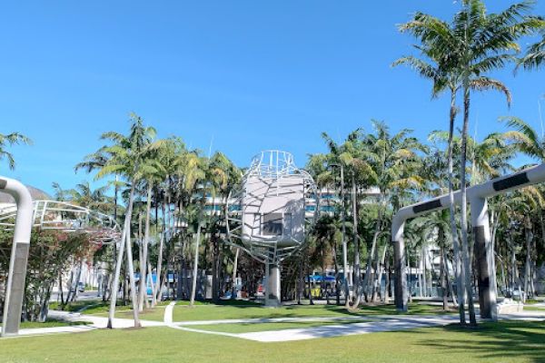 A park with palm trees, modern sculptures, and a building in the background under a clear blue sky.