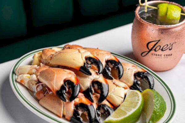 A plate of stone crab claws with lime wedges and sauce, accompanied by a drink in a branded cup on the side of the table.