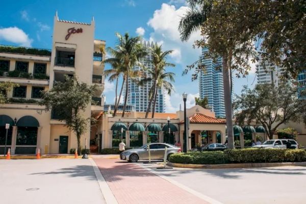 The image shows a sunny street scene with a restaurant, palm trees, and modern buildings in the background, under a clear blue sky.