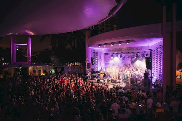 A large crowd enjoys a live concert in an outdoor venue with vibrant stage lighting and performers on stage.