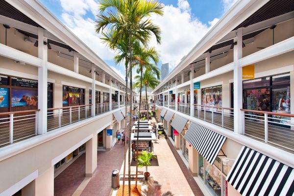 An outdoor shopping mall with two levels of shops, striped awnings, palm trees, and people walking.