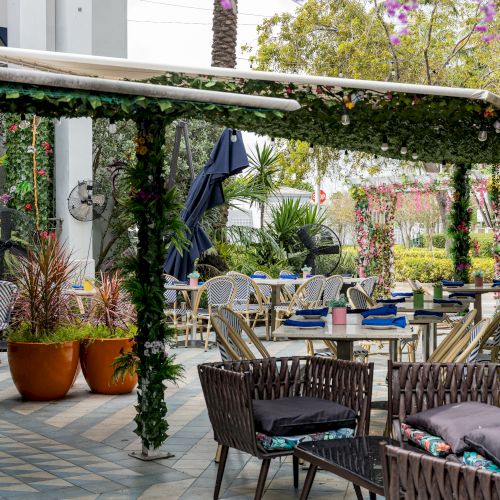 An outdoor dining area with tables and chairs, decorated with vibrant plants and flowers, under a partially covered pergola.