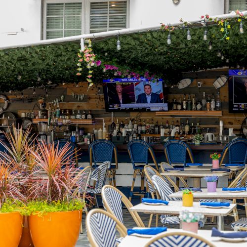 Outdoor bar with tables, blue chairs, and hanging greenery. TVs display sports news. Orange planters hold decorative plants.