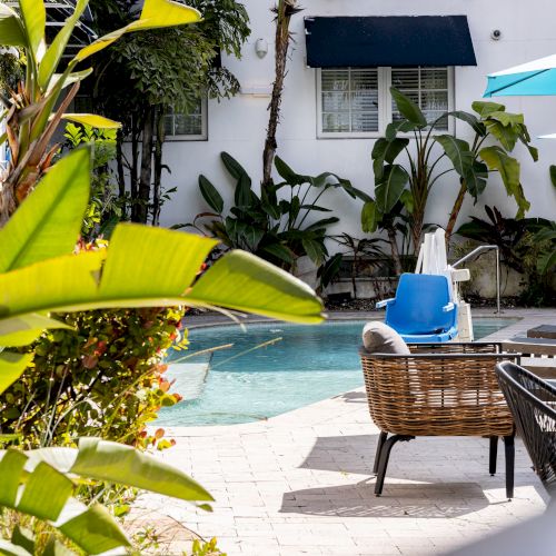A serene poolside scene with tropical plants, lounge chairs, and a blue umbrella under sunny skies.