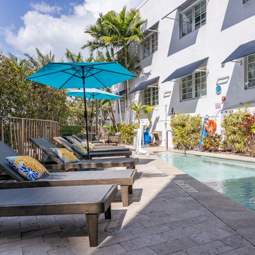 A sunny poolside scene with lounge chairs, blue umbrellas, and decorative pillows, surrounded by greenery against a white building.