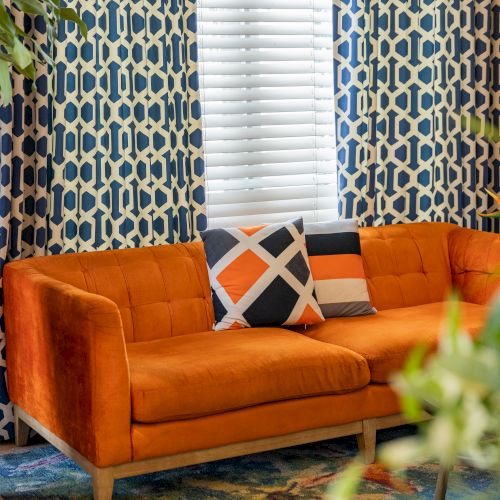 An orange couch with patterned pillows is in front of blue geometric curtains and a colorful rug, near a window with blinds.