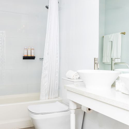 White bathroom with a bathtub, shower curtain, toilet, towels, toiletries, and a vessel sink on a countertop.