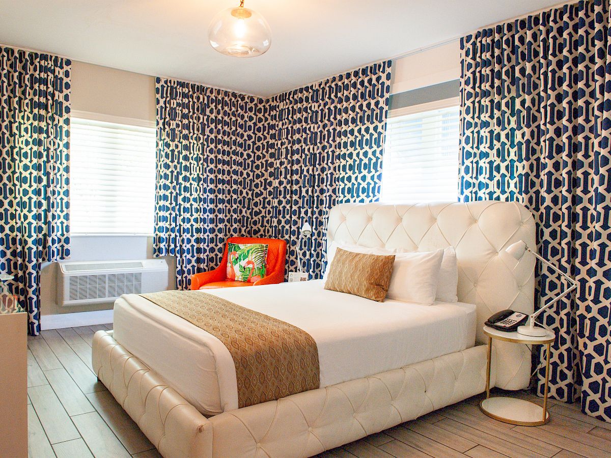 A stylish bedroom with a white bed, geometric blue curtains, an orange chair, and modern lighting, exuding comfort and elegance.