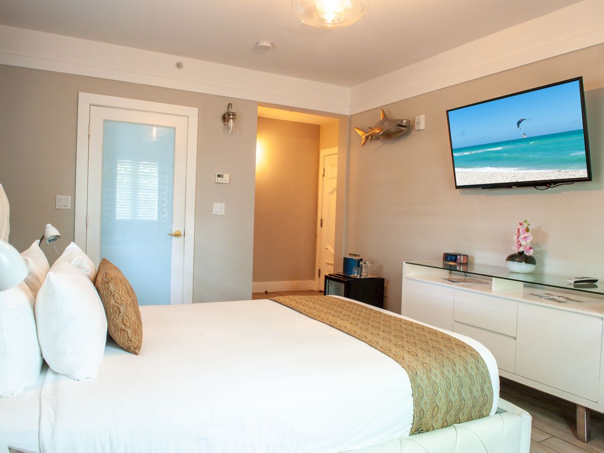 A hotel room with a bed, pillows, a TV displaying a beach scene, a decorative shark, a dresser, and a vase with flowers on top.