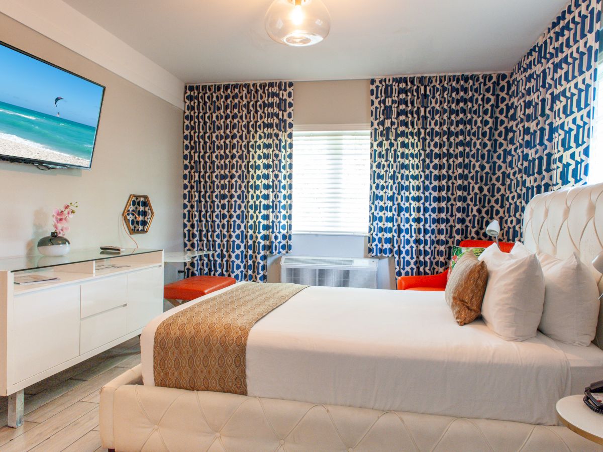 The image shows a modern bedroom with a white bed, blue patterned curtains, a wall-mounted TV, and a sideboard with a vase.