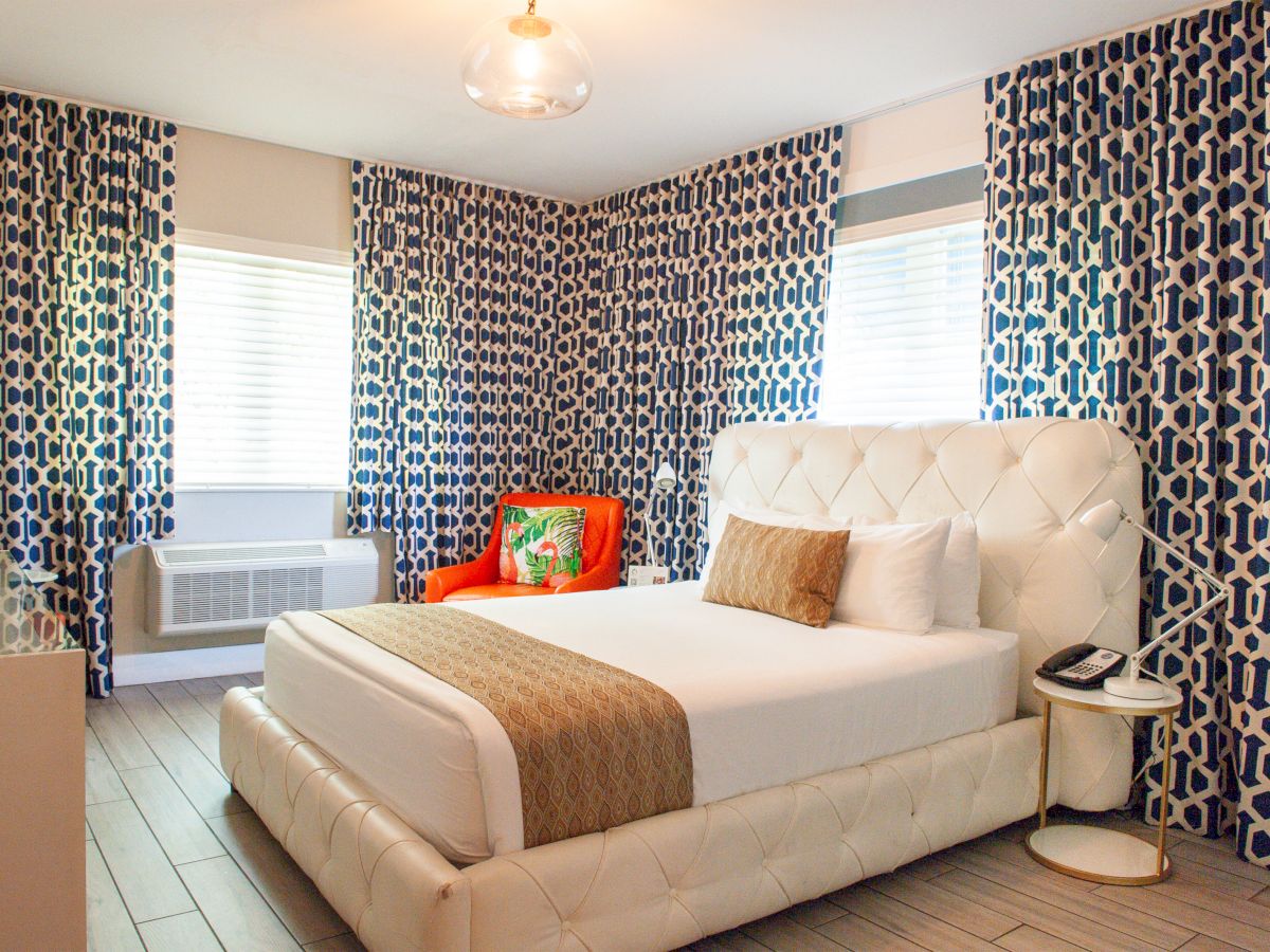 A modern bedroom with a white bed, geometric-patterned curtains, an orange chair, and a small side table with a lamp and phone.