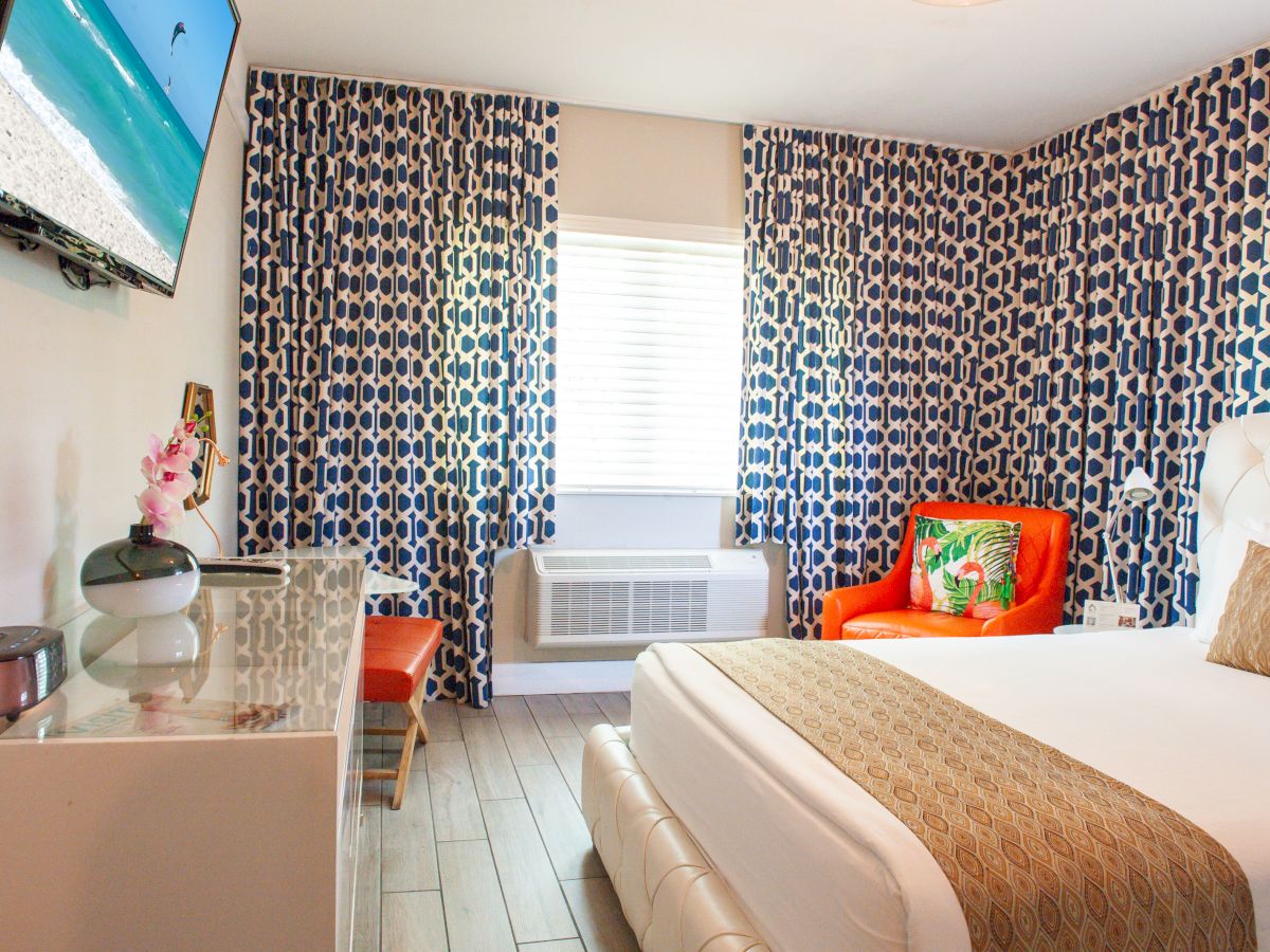 A cozy hotel room with a patterned blue curtain, TV, orange chair, bed, and decor. Bright and modern with natural light filling the space.