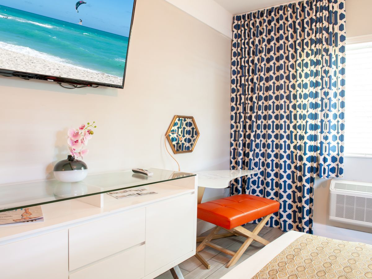 A room with a TV showing a beach scene, geometric blue curtains, orange stool, small desk, hexagonal mirror, and a vase with flowers.
