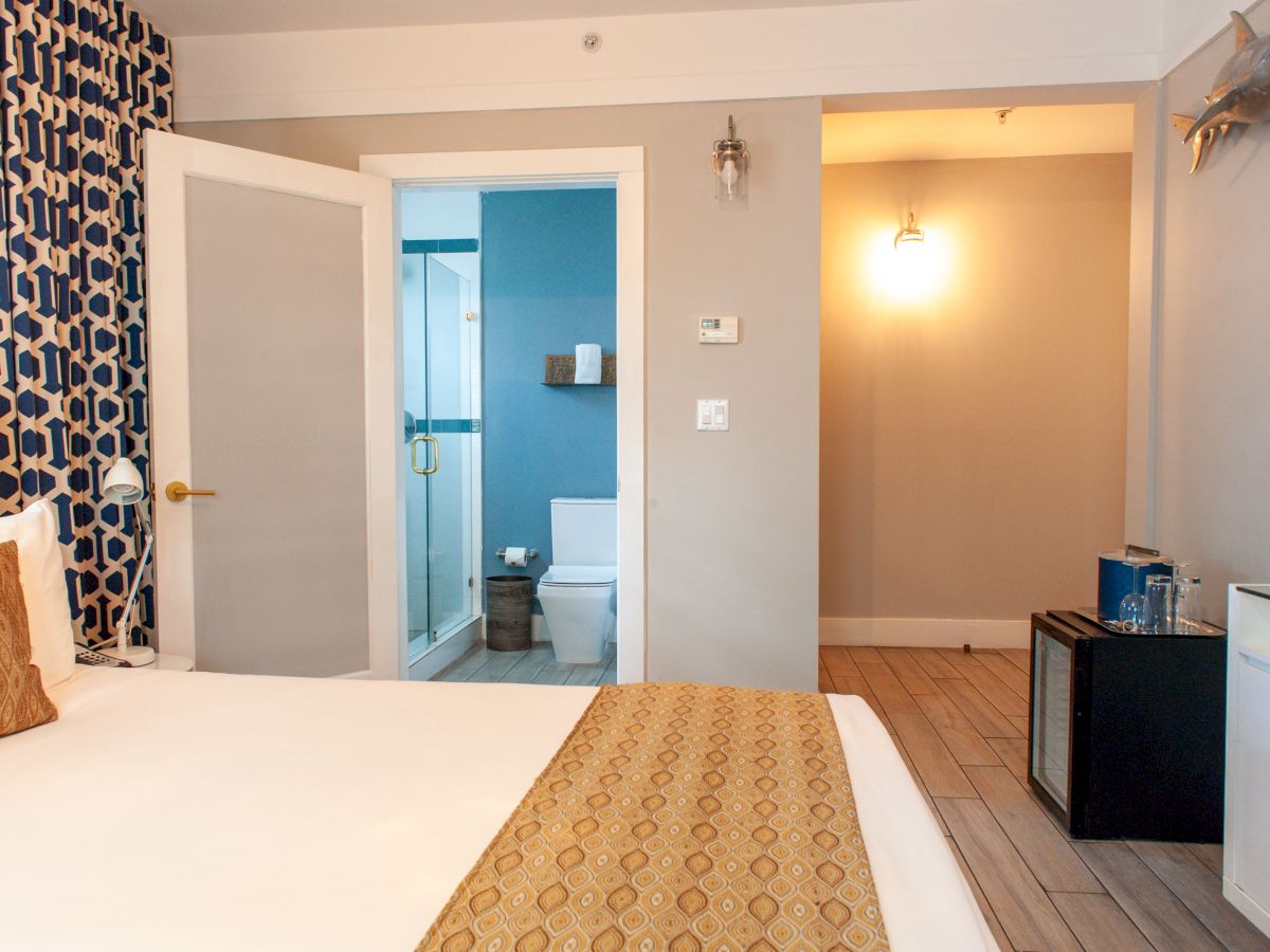 A modern hotel room featuring a bed, patterned wallpaper, an open bathroom door, a mini fridge, and wall-mounted lights.