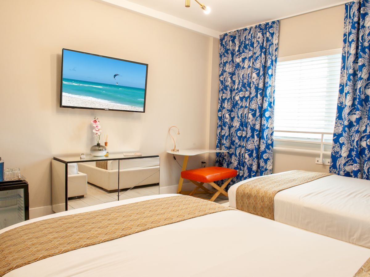 A hotel room with two beds, a wall-mounted TV showing a beach, blue patterned curtains, a desk, small fridge, and decorative elements visible.