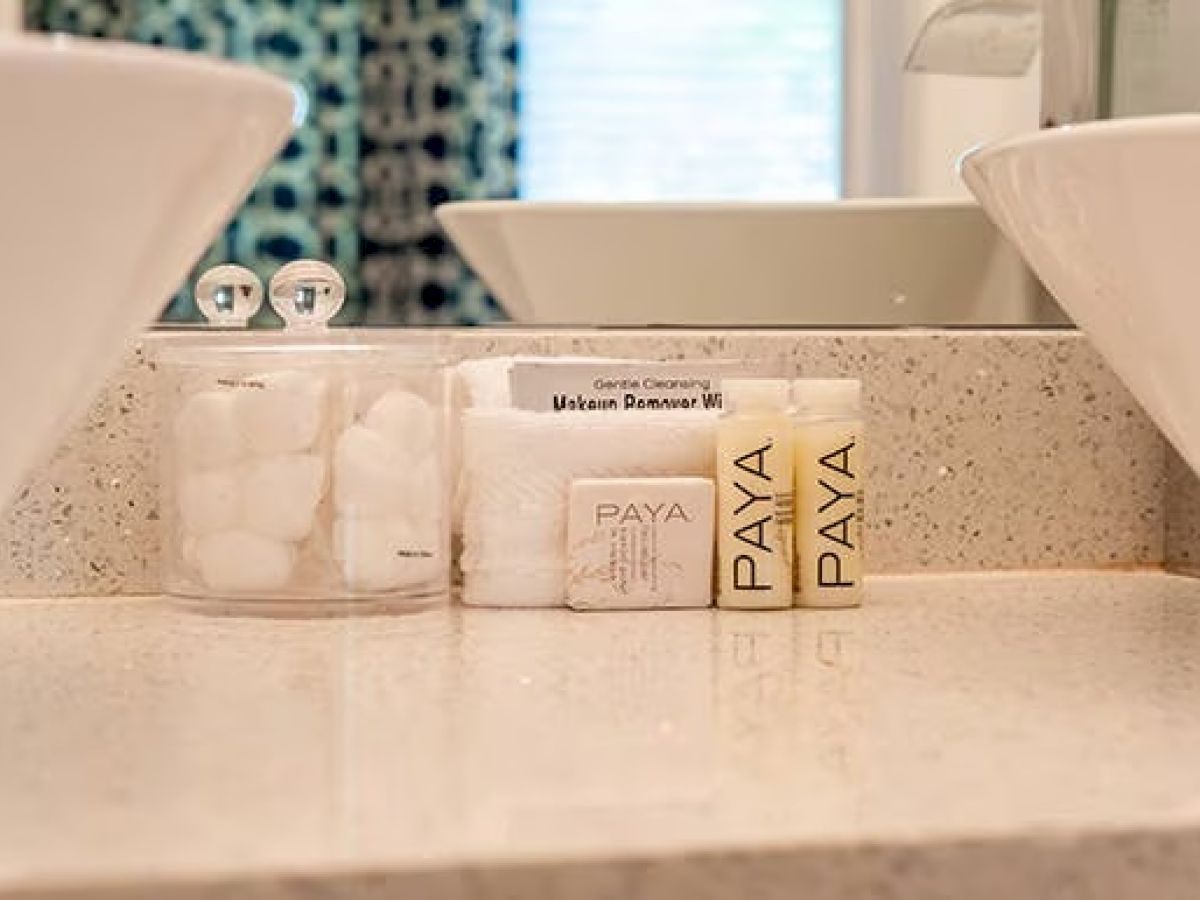The image shows a bathroom countertop with a container of cotton balls, Paya toiletries, and makeup remover wipes, near a sink.