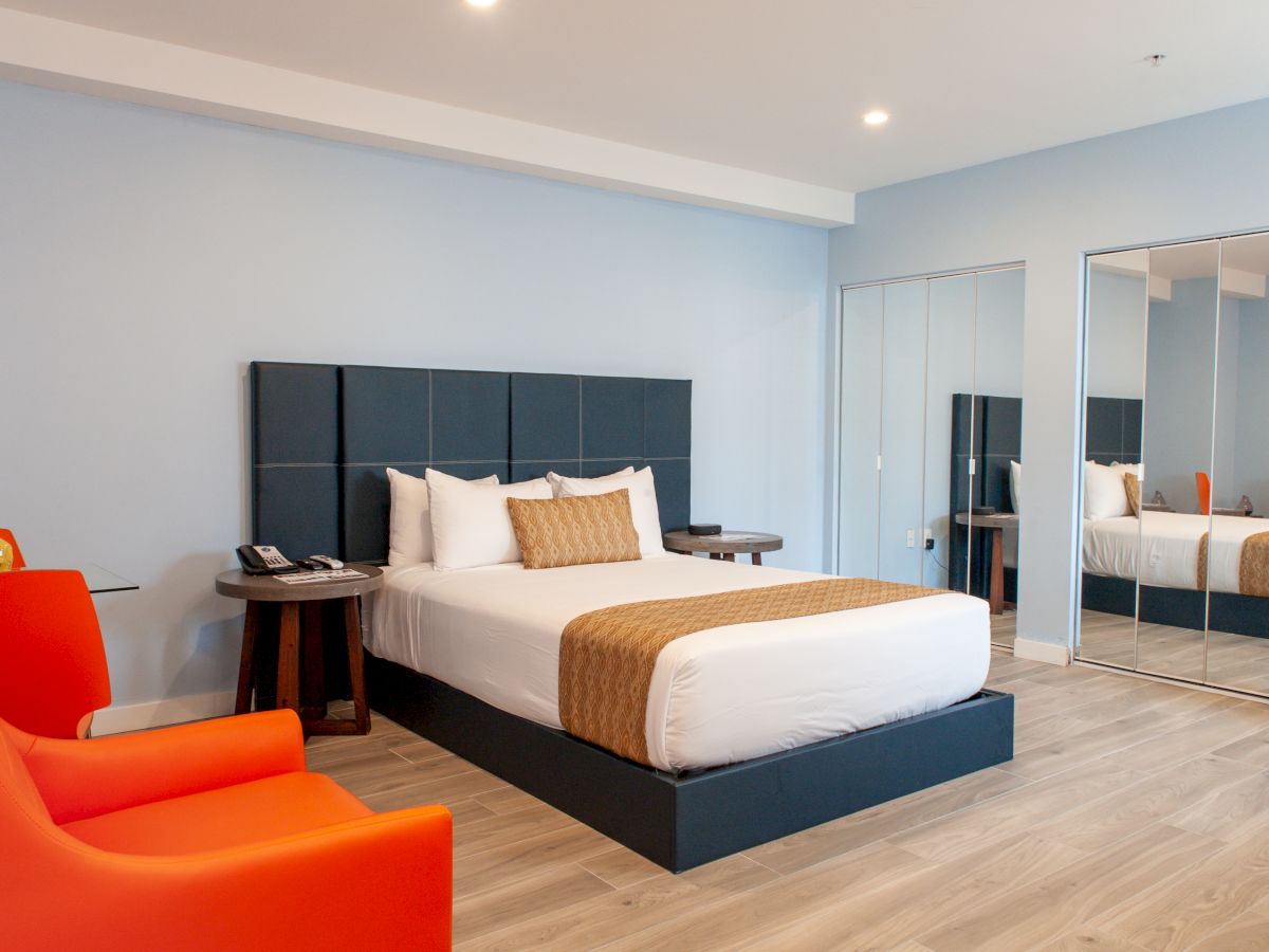 A modern hotel room with a large bed, patterned pillows, orange chairs, and mirrored closet doors.