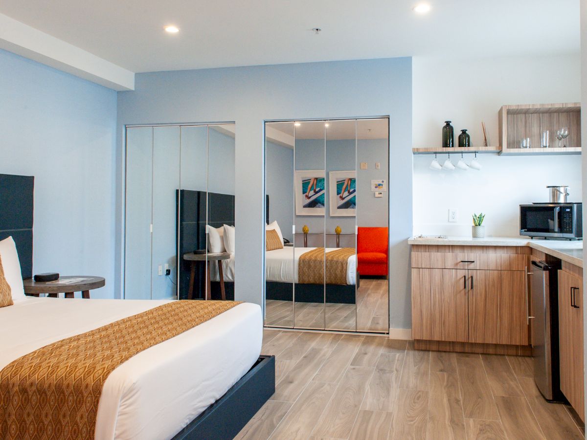 Modern hotel room with a bed, mirrored wardrobe, kitchenette, and decor in neutral tones, offering a comfortable stay.