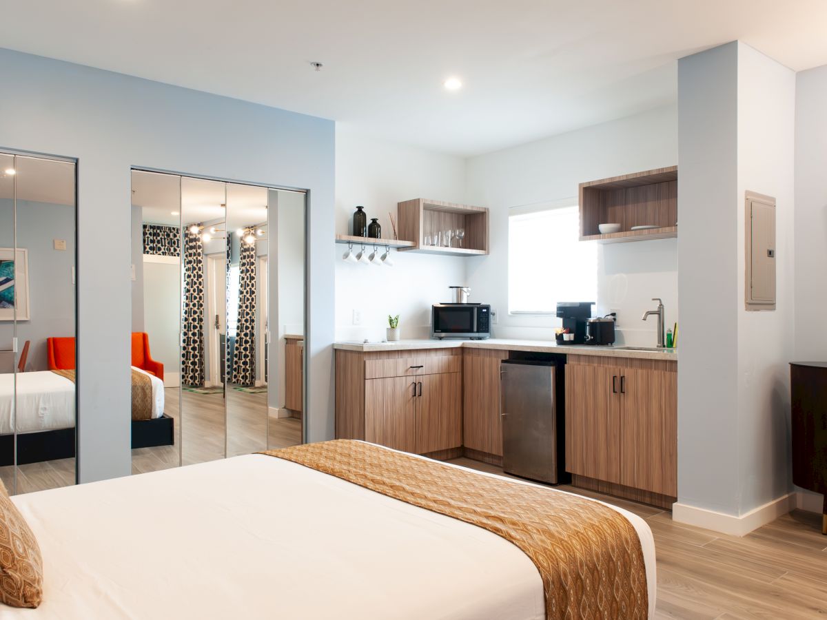 A modern hotel room with a bed, kitchenette, TV, and mirrored closet doors. Bright and neatly arranged space for a comfortable stay.