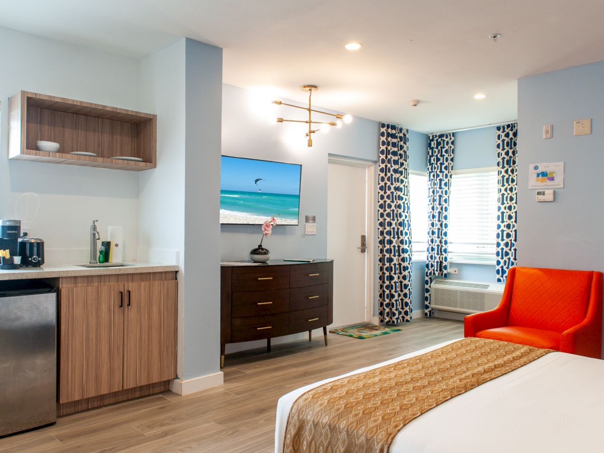 The image shows a modern hotel room with a bed, kitchenette, dresser, TV, orange chair, and patterned curtains.
