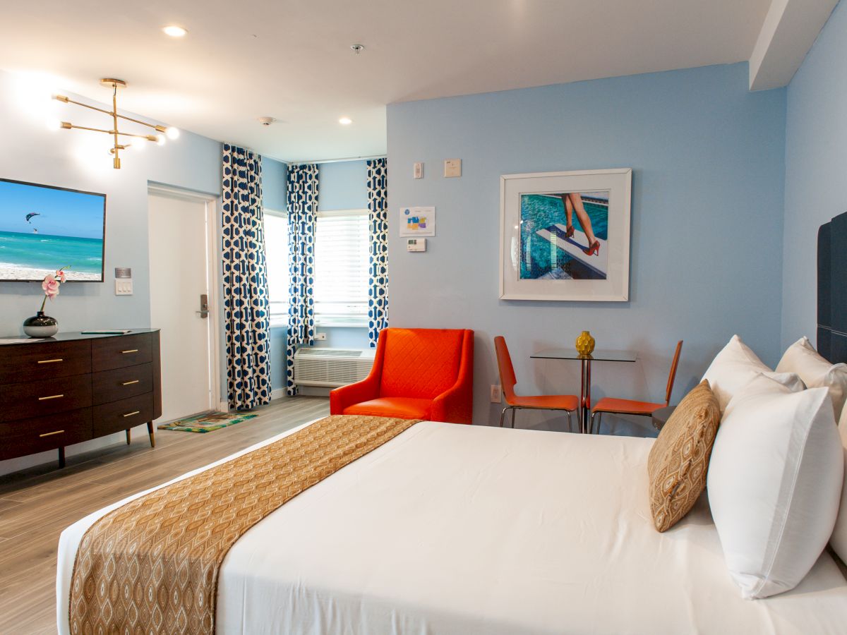 A modern hotel room with a TV, dresser, orange chair, small table, and bed, decorated in blue tones with geometric curtains.