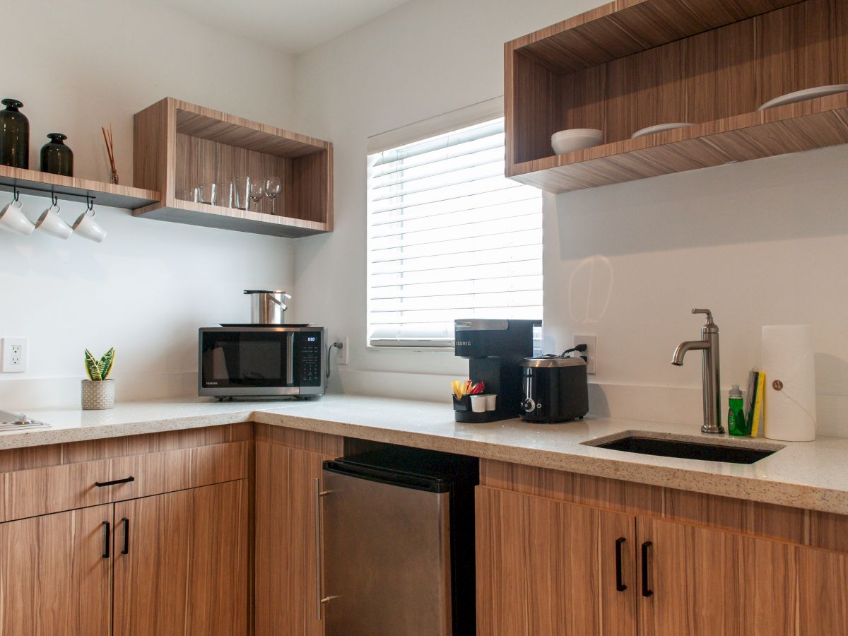A modern kitchen with wood cabinets, a microwave, a coffee maker, a toaster, a sink, and shelves with mugs and glassware.