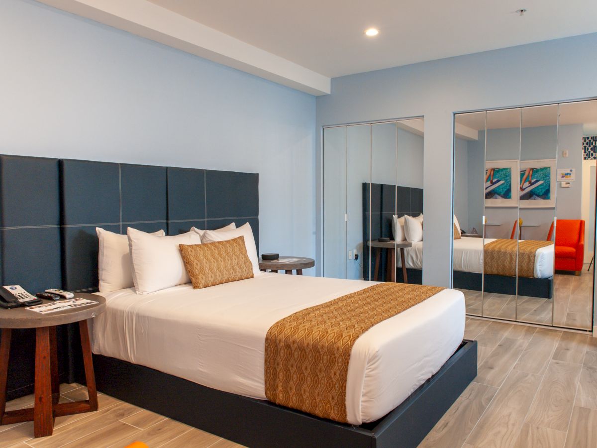 A modern hotel room with a bed, decorative pillows, mirrored closet, and an orange chair in a clean, contemporary setting.