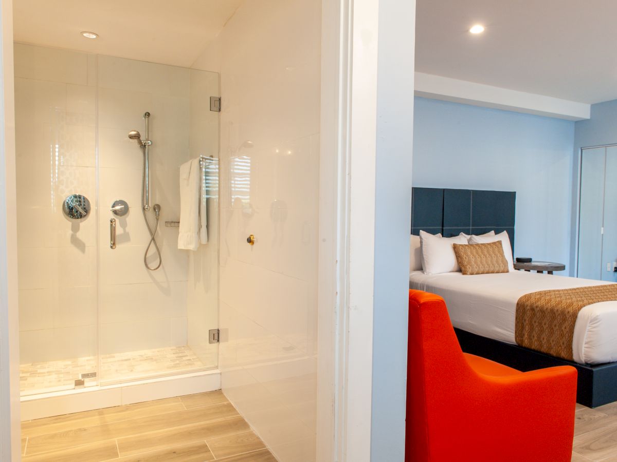 A modern hotel room with a glass shower, wooden floors, a bed, and an orange chair, creating a bright and cozy atmosphere.