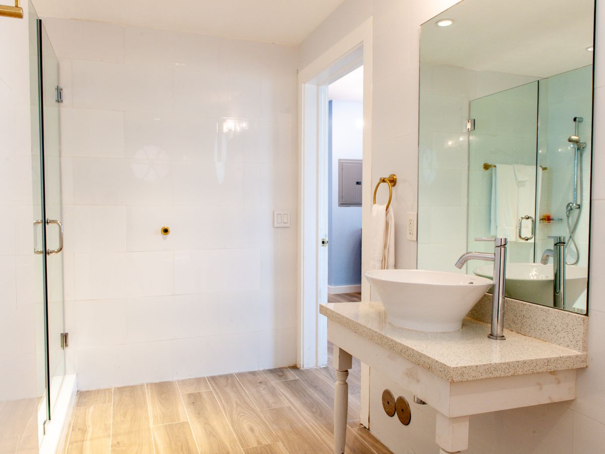 A modern bathroom with a glass shower, a vessel sink on a countertop, a large mirror, and wooden flooring.