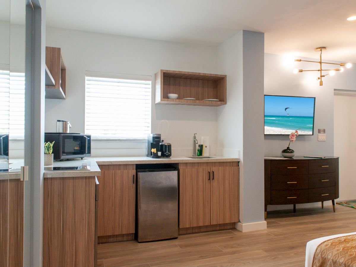 The image shows a modern kitchen with wooden cabinets, a microwave, and a small fridge; a TV and a dresser are visible in the room.