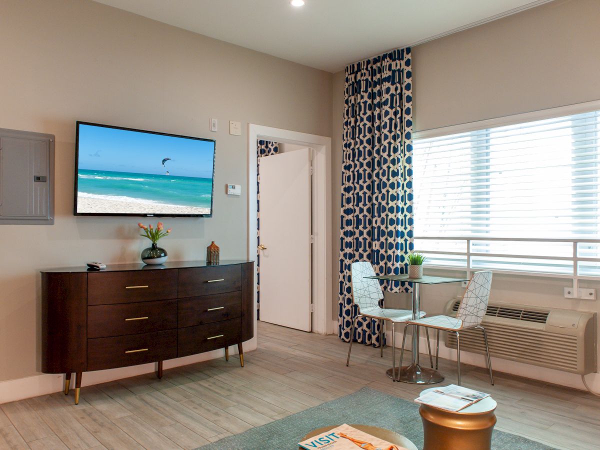 A furnished room with a TV showing a beach, a dresser, a small table with two chairs, patterned curtains, and a window with blinds.