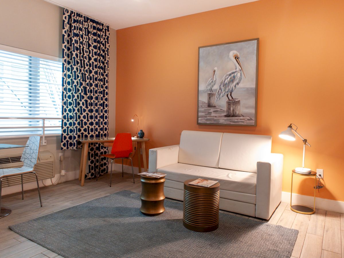 A cozy living room with an orange wall, white sofa, and two pelicans painting. Round tables, floor lamp, and a desk with an orange chair.