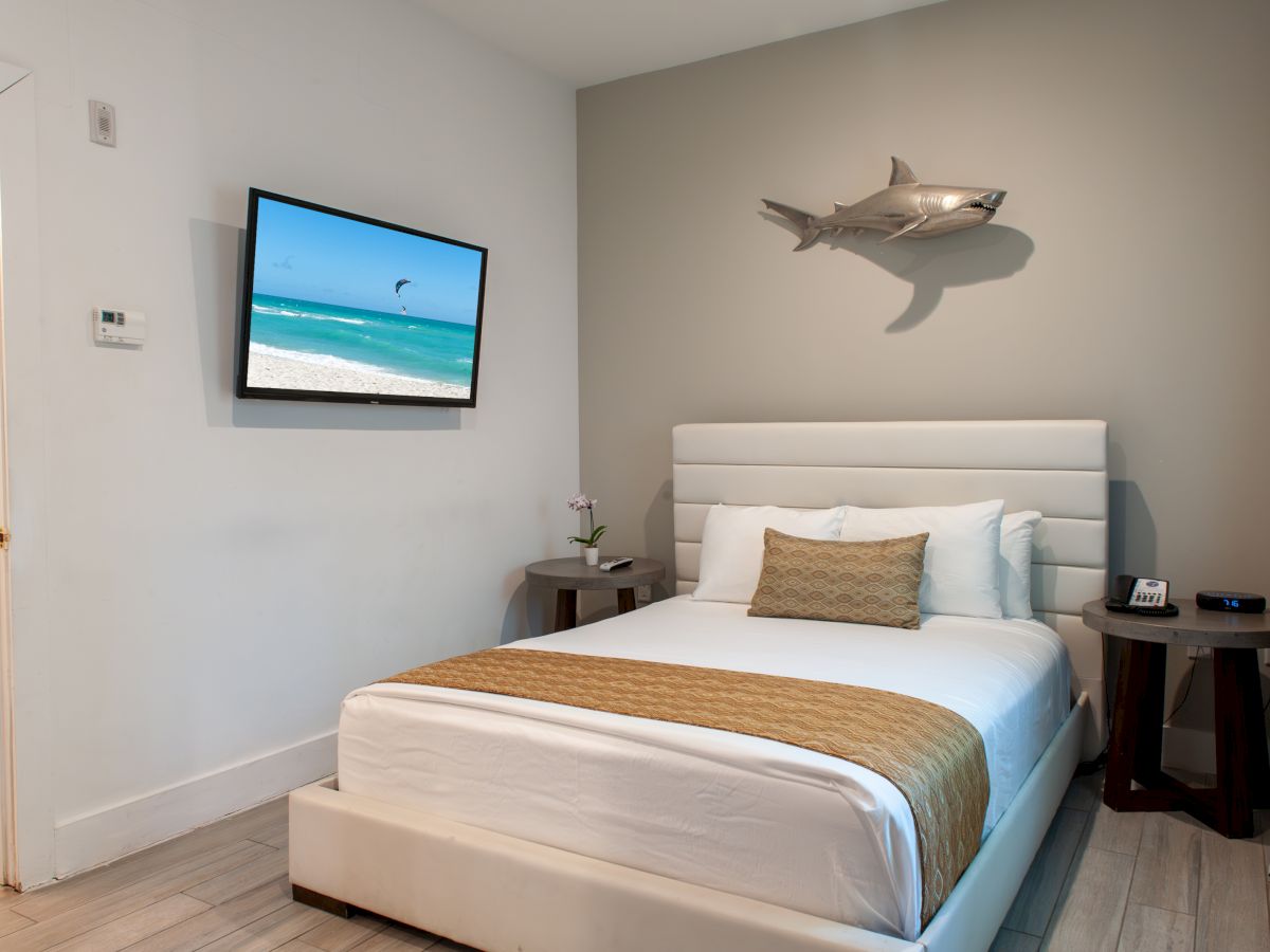 A modern bedroom with a bed, TV showing a beach scene, shark wall decor, nightstands, and a digital clock at 7:16.