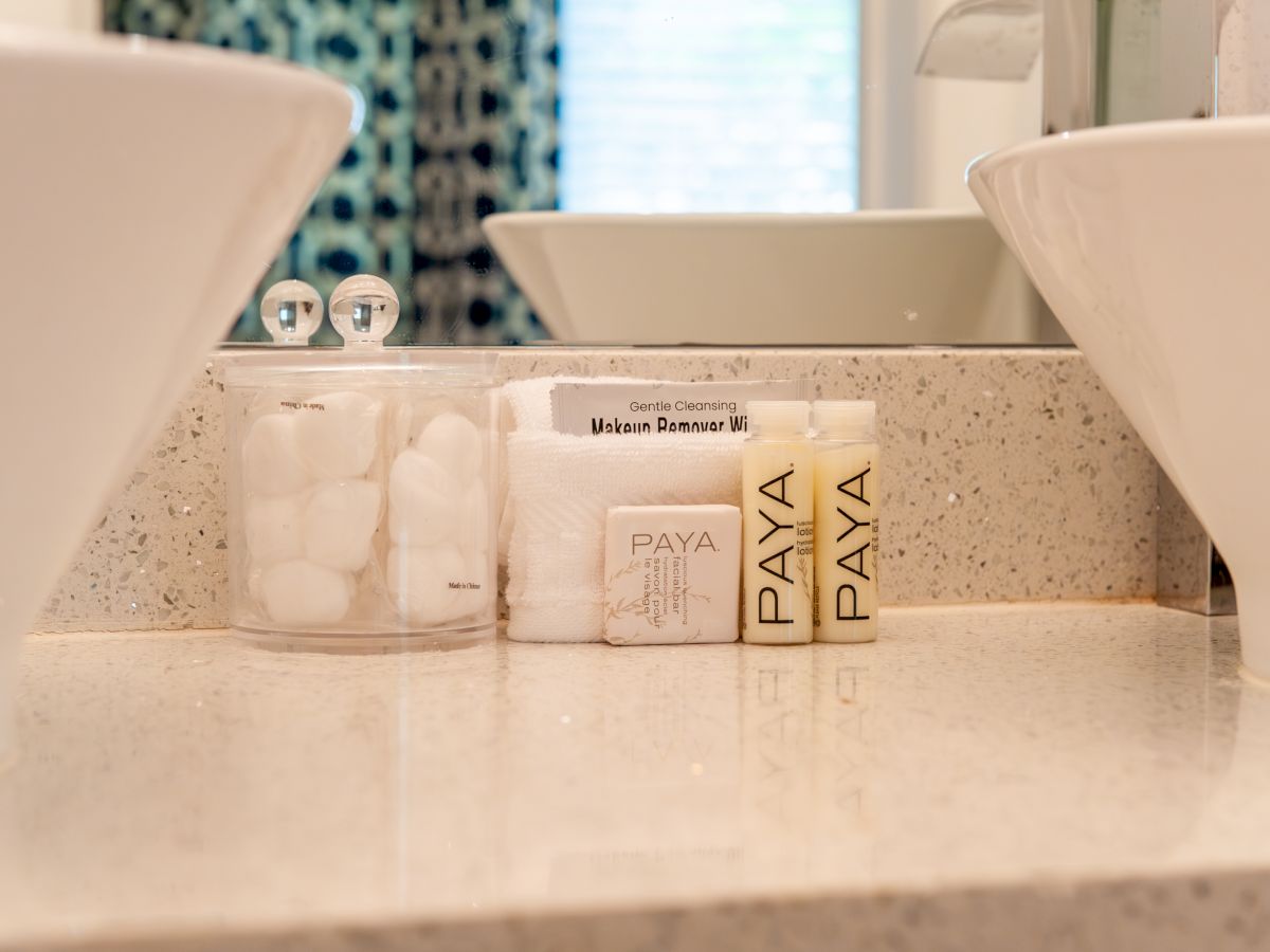 A bathroom counter with cotton balls, makeup remover wipes, soap, and Paya brand lotion bottles, next to a sink.
