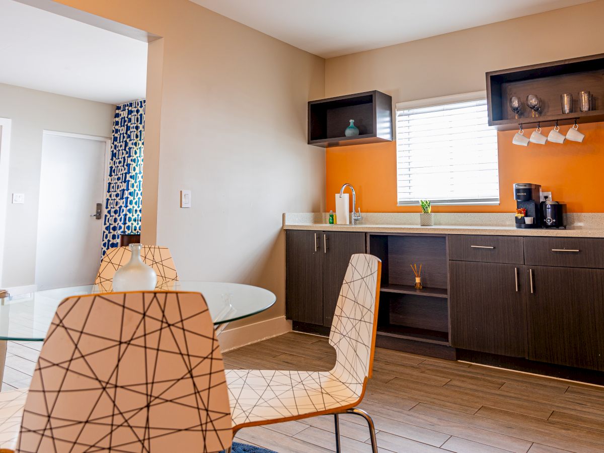 A modern kitchen with a sink, coffee maker, geometric chairs, and a glass table, featuring a blue patterned curtain and orange accent wall.