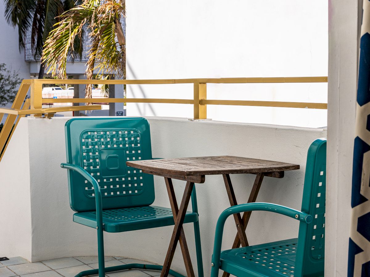 A small patio with two teal chairs and a wooden table, surrounded by white walls and yellow railing in a sunny setting.