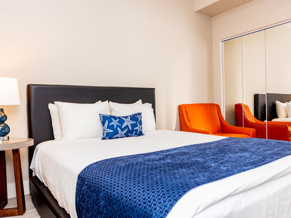 A bedroom with a bed, blue starfish pillow, orange chair, side table with a lamp, and mirrored closet doors.