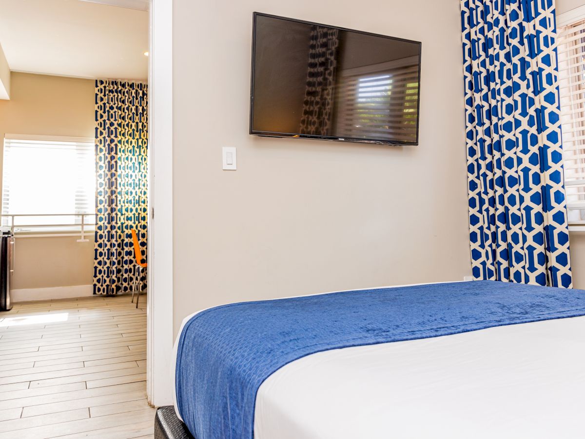 A room with a bed, TV, patterned curtains, window blinds, and a view into a kitchen area with tiled flooring.