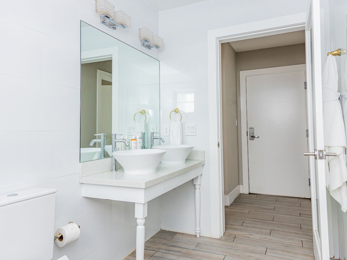A modern bathroom with dual sinks, large mirror, white towels, and two lights. A toilet paper holder and a door are also visible.