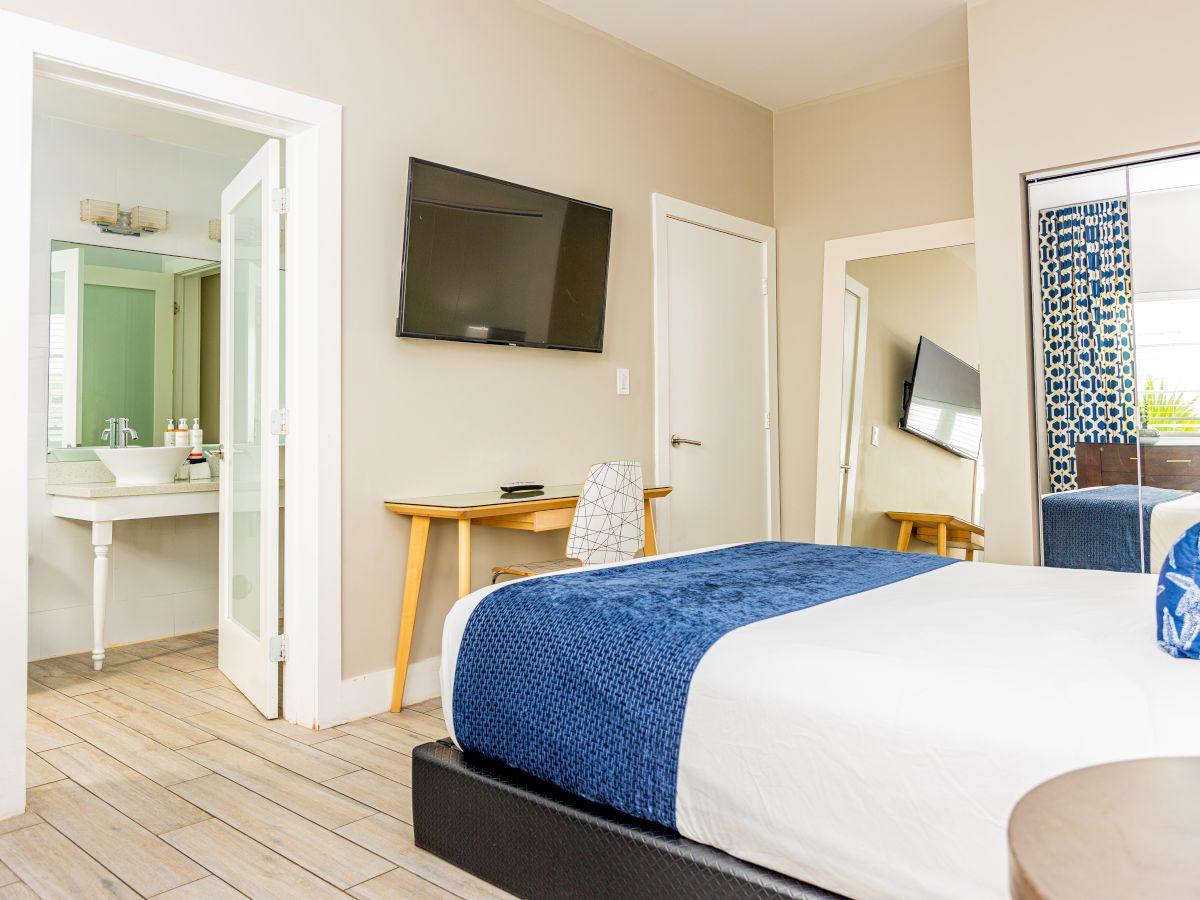 A cozy hotel room with a bed, TV, desk, chair, mirror, and an open door to the bathroom. The decor is modern and inviting.
