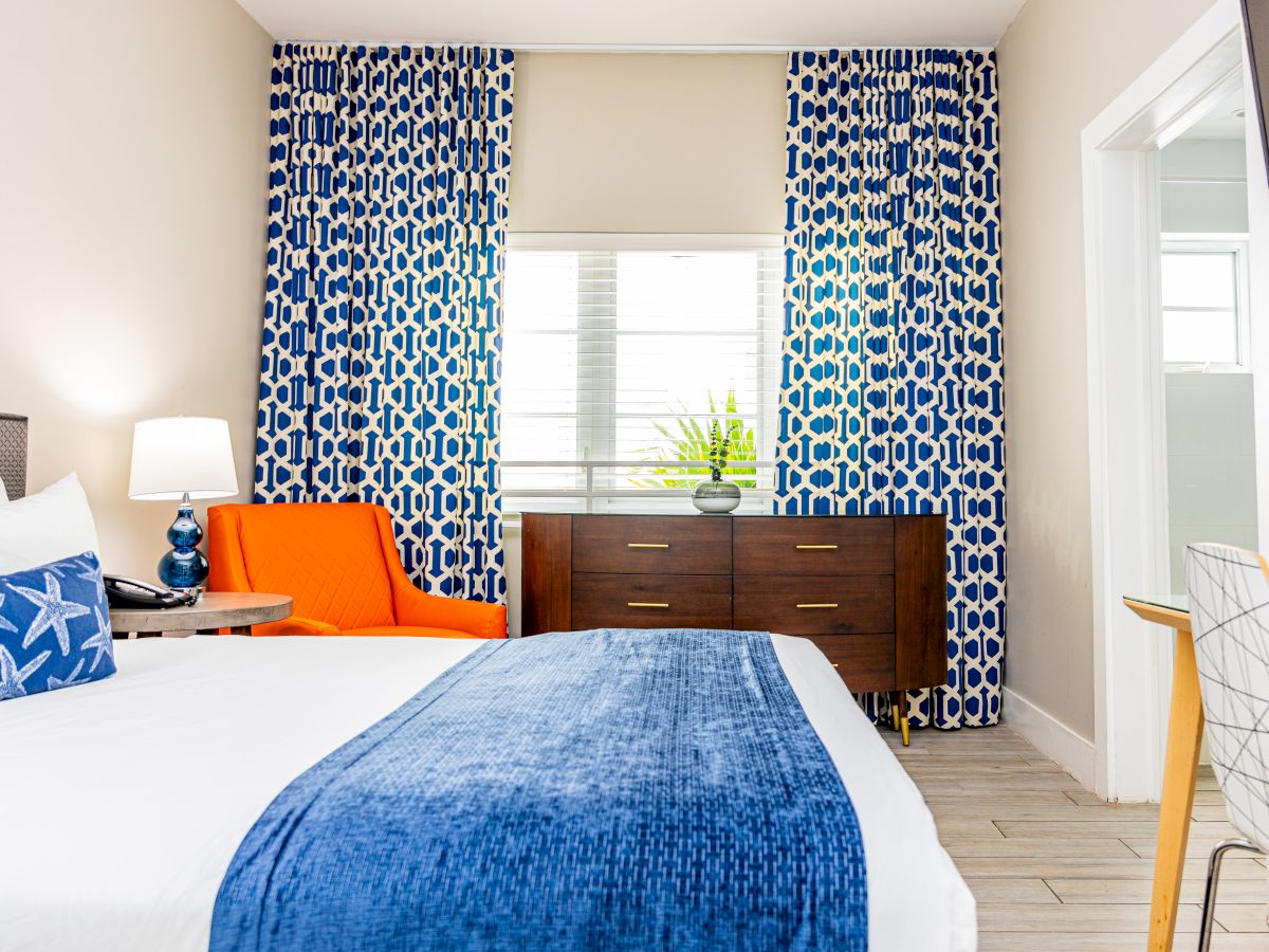 A bright bedroom with a blue and white theme, featuring an orange chair, blue patterned curtains, and a wooden dresser.