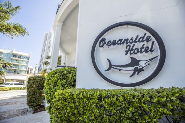 Sign for "Oceanside Hotel" with a swordfish logo, surrounded by greenery and nearby modern buildings under a clear sky.